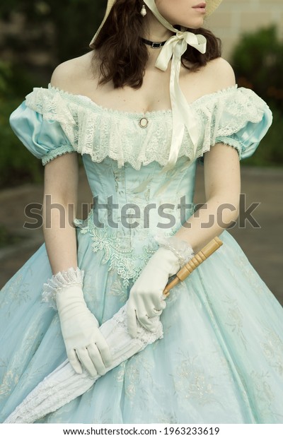 A beautiful young woman in a historical\
blue dress with an umbrella, gloves and a hat. Dress on a woman of\
the 19th century. Design for the book\
cover