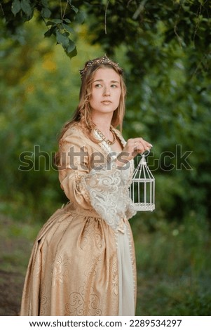 A beautiful young woman in a historical golden dress with a crown on her head holds a white cage in her hands. Princess in a medieval dress in the forest. queen in the garden