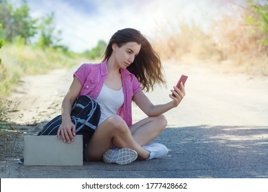 Beautiful Young Woman Hitchhiker With A Backpack Holds A Cardboard With A Place For Text Sitting On The Road And Looks Into The Phone. Hitchhiking With A Mobile Cell Phone. Copyspace