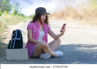 Beautiful Young Woman Hitchhiker In A Cap With A Backpack And A Cardboard With A Place For Text Sits On The Road And Looks Into The Phone. Hitchhiking With A Mobile Cell Phone. Copyspace