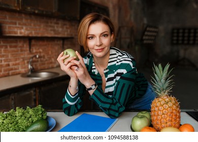 Beautiful Young Woman Holding Apple And Looking To Camera In Her Modern Loft Kitchen. Fitness Coach. Healthy Food Concept.