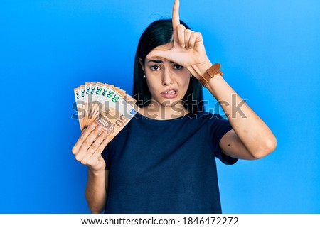 Beautiful young woman holding bunch of 50 euro banknotes making fun of people with fingers on forehead doing loser gesture mocking and insulting. 