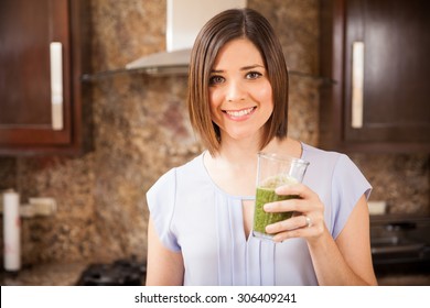 Beautiful young woman holding a glass of green and healthy juice at home and smiling - Powered by Shutterstock