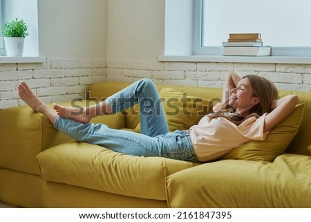 Beautiful young woman holding hands behind head and smiling while relaxing on the couch at home