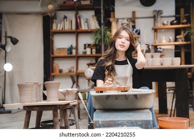 Beautiful young woman holding pottery instrument for scraping, smoothing, shaping and sculpting. Lady siting on bench with pottery wheel and making clay pot - Powered by Shutterstock