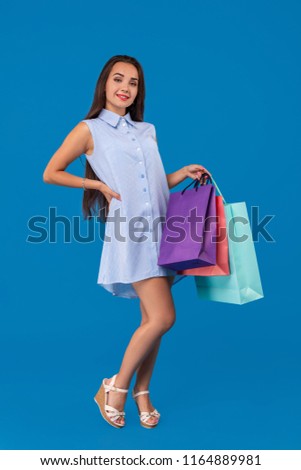 Beautiful young woman holding shopping bags and looking at the camera while standing against blue background.