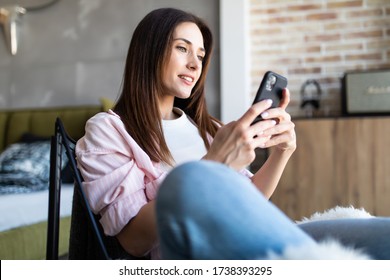 Beautiful Young Woman Holding Smart Phone And Looking At Camera With Smile While Sitting In Big Comfortable Chair At Home