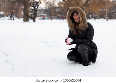 Beautiful Young Woman Holding Snowball, Wearing Fur Hooded Jacket And Smiling. Snow All Around Her. 