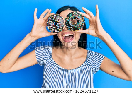 Beautiful young woman holding tasty colorful doughnuts on eyes smiling and laughing hard out loud because funny crazy joke. 