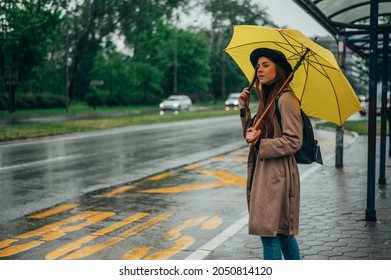 Beautiful young woman holding yellow umbrella a waiting for a bus on a bus stop on a rainy day