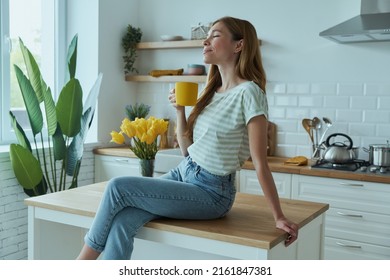 Beautiful young woman holding yellow cup and smiling while sitting on the kitchen counter