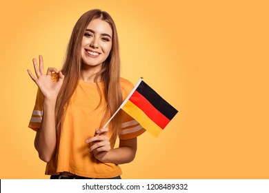 A beautiful young woman holds in her hands the flag of Germany and shows the OK sign. Exchange student, learn Germanic language. Tourist traveling. Yellow, orange background. Football fan.