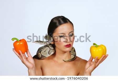 A beautiful young woman holds yellow and orange 
peppers in her hands while a python snake curls 
itself around her.