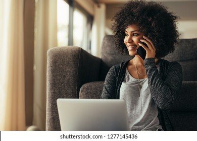 Beautiful young woman at home with laptop and talking on cell phone and smiling. Female using cell mobile phone while sitting at home.