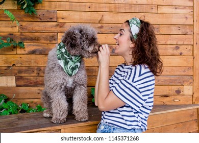 Beautiful young woman hugging her dog, brown Spanish water dog over wood background. Daytime, love for animals concept and lifestyle. Dog wearing a green leaves bandana, woman wearing casual clothes
