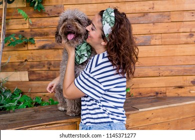 Beautiful young woman hugging her dog, brown Spanish water dog over wood background. Daytime, love for animals concept and lifestyle. Dog wearing a green leaves bandana, woman wearing casual clothes