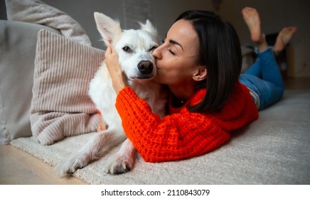 Beautiful young woman is hugging and kissing her big white cute dog while lying on the carpet on home floor
