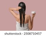 Beautiful young woman with jar of cosmetic product applying hair mask on pink background, back view