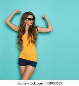Beautiful young woman in jeans shorts and orange shirt posing with arms raised. Three quarter length studio shot on teal background.