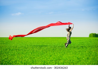 Beautiful Young Woman Jumping On A Green Meadow With A Colored Red Tissue. Happy Female In Wheat Field With Fabric. Summer Picnic And Freedom Concept.