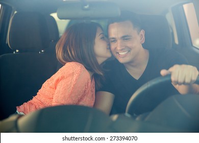 Beautiful young woman kissing her boyfriend in the cheek while he drives. Shot through the windshield