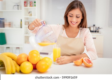 Beautiful young woman in the kitchen squeezing orange juice with an electric juicer. She is pouring juice into a glass. - Powered by Shutterstock