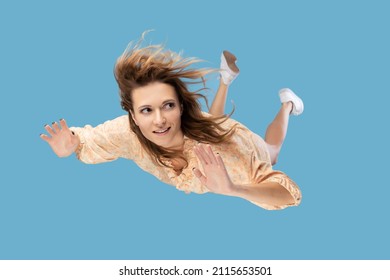 Beautiful young woman levitating in mid-air, falling down and her hair messed up soaring from wind, model flying hovering with dreamy peaceful expression. indoor shot isolated on blue background