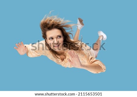 Beautiful young woman levitating in mid-air, falling down and her hair messed up soaring from wind, model flying hovering with dreamy peaceful expression. indoor shot isolated on blue background