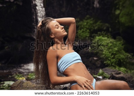 Beautiful young woman in light blue bikini relaxing near mountain waterfall
