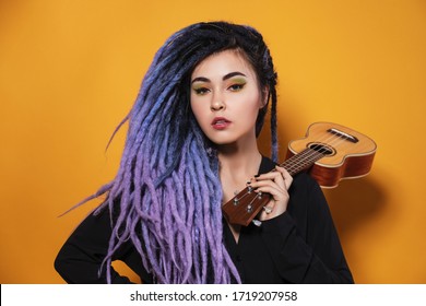 beautiful young woman with lilac dreadlocks and multi-colored makeup stands on a yellow studio background and holds an ukulele on her shoulder