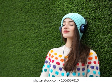 Beautiful young woman listening to music with headphones near grass wall. Space for text