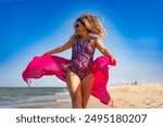 Beautiful young woman with long blond hair in colorful pink swimsuit and sunglasses walking with pink scarf under arms fluttering strongly in wind on sandy beach on sunny and windy day. Front view