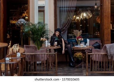 Beautiful young woman with long brunette hair in black beret and leather jacket sitting in Paris street cafe - Shutterstock ID 1570010605