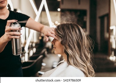 Beautiful young woman with long curly hair in hair salon. Professional hairdresser styling with hairspray.
