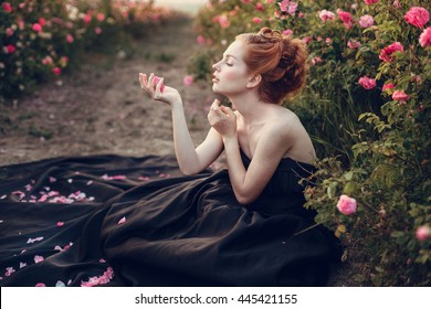 Beautiful young woman with long curly hair posing near roses in a garden. The concept of perfume advertising. 