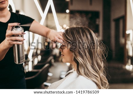 Beautiful young woman with long curly hair in hair salon. Professional hairdresser styling with hairspray.