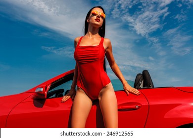 schöne junge Frau mit langen dunklen Haaren in rotem KarosAnzug und Sonnenbrille, die mit rotem Cabrio steht