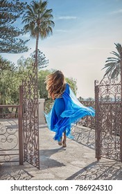 Beautiful Young Woman With Long Dark Hair Wearing Long Blue Dress And Running Away On Stunning Sea View Background At Summer 