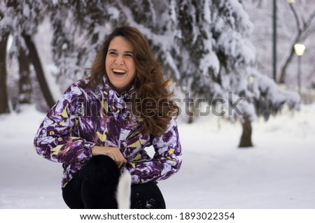 A beautiful young woman with long hair in a winter snowy park is not posing but laughing frankly happily .. Winter portrait of a woman with brown hair among the snow.
