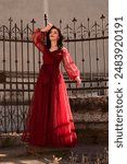 Beautiful young woman in a long red gown pretending to faint princess style in front of a gothic wrought iron gate