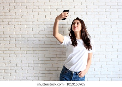 Beautiful Young Woman, Long Wavy Brunette Hair, Dressed In 90s Style, Making Selfie. Female Wearing Mom Jeans & Plain White T Shirt Taking Pictures Of Herself. White Brick Wall Background, Copy Space.