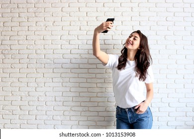 Beautiful Young Woman, Long Wavy Brunette Hair, Dressed In 90s Style, Making Selfie. Female Wearing Mom Jeans & Plain White T Shirt Taking Pictures Of Herself. White Brick Wall Background, Copy Space.