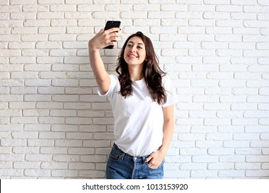 Beautiful young woman, long wavy brunette hair, dressed in 90s style, making selfie. Female wearing mom jeans & plain white t shirt taking pictures of herself. White brick wall background, copy space.