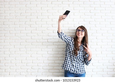 Beautiful Young Woman, Long Wavy Brunette Hair, Dressed In 90s Style, Making Selfie. Female Wearing Mom Jeans & Checkered Shirt Taking Pictures Of Herself. White Brick Wall Background, Copy Space.