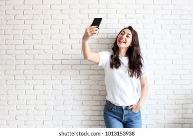 Beautiful Young Woman, Long Wavy Brunette Hair, Dressed In 90s Style, Making Selfie. Female Wearing Mom Jeans & Plain White T Shirt Taking Pictures Of Herself. White Brick Wall Background, Copy Space.