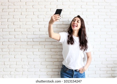 Beautiful Young Woman, Long Wavy Brunette Hair, Dressed In 90s Style, Making Selfie. Female Wearing Mom Jeans & Plain White T Shirt Taking Pictures Of Herself. White Brick Wall Background, Copy Space.