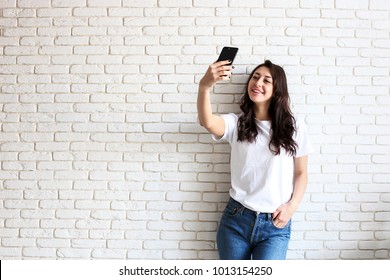 Beautiful Young Woman, Long Wavy Brunette Hair, Dressed In 90s Style, Making Selfie. Female Wearing Mom Jeans & Plain White T Shirt Taking Pictures Of Herself. White Brick Wall Background, Copy Space.