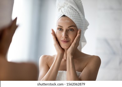 Beautiful young woman look in mirror massaging face applying cream in bathroom, pretty lady wrap towel on head put facial moisturizer lifting hydrating moisturizing creme, skin care treatment concept
