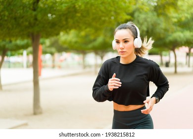 Beautiful Young Woman Looking Focused While Running Fast In The Park. Hispanic Woman In Her 30s Listening To Music With Headphones Working Out Outdoors