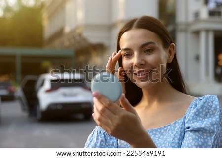 Beautiful young woman looking at herself in cosmetic pocket mirror outdoors. Space for text